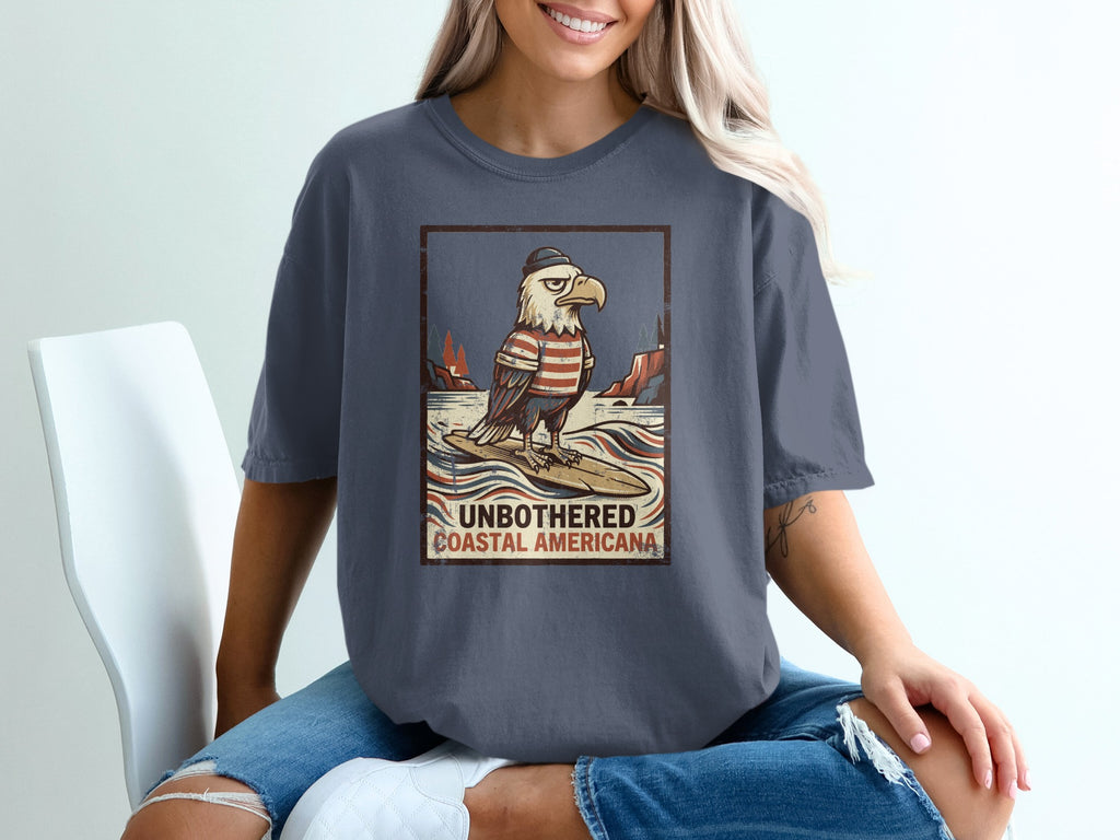 T-shirt graphic of an eagle surfing with text "UNBOTHERED COASTAL AMERICANA".