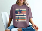 Purple t-shirt with silhouetted palm trees and sailboat on striped background.