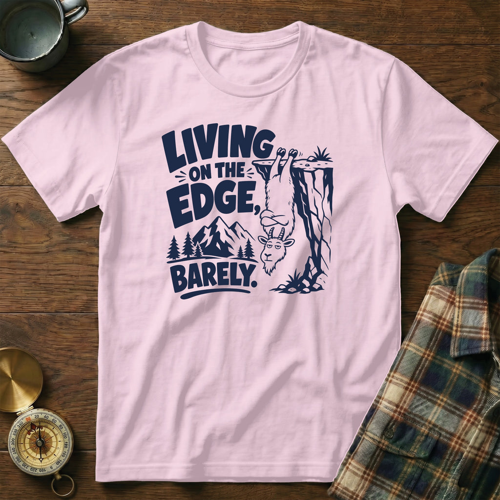 Pink t-shirt with navy graphic of a goat hanging from a cliff, text "LIVING ON THE EDGE, BARELY."