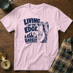 Pink t-shirt with navy graphic of a goat hanging from a cliff, text "LIVING ON THE EDGE, BARELY."