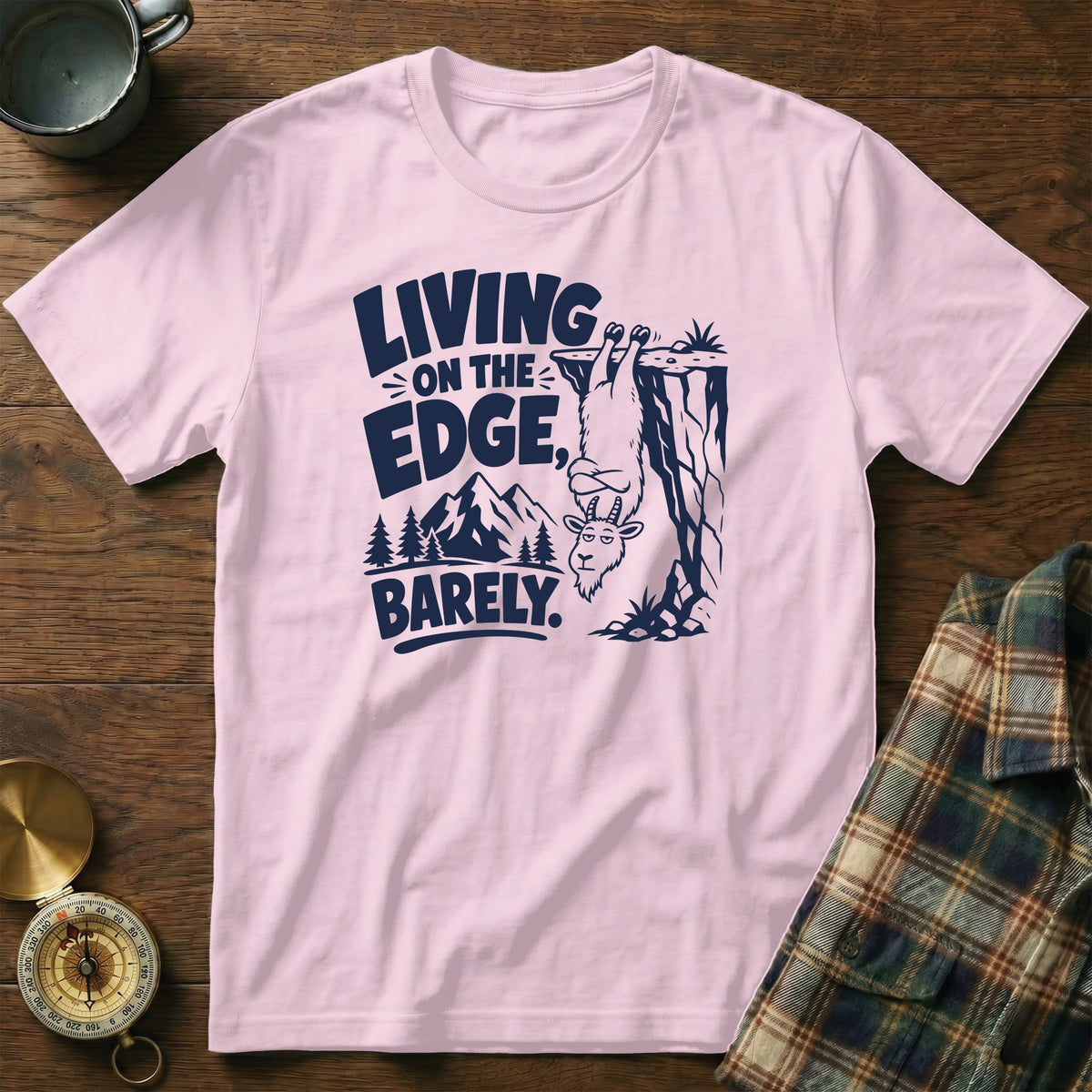 Pink t-shirt with navy graphic of a goat hanging from a cliff, text "LIVING ON THE EDGE, BARELY."