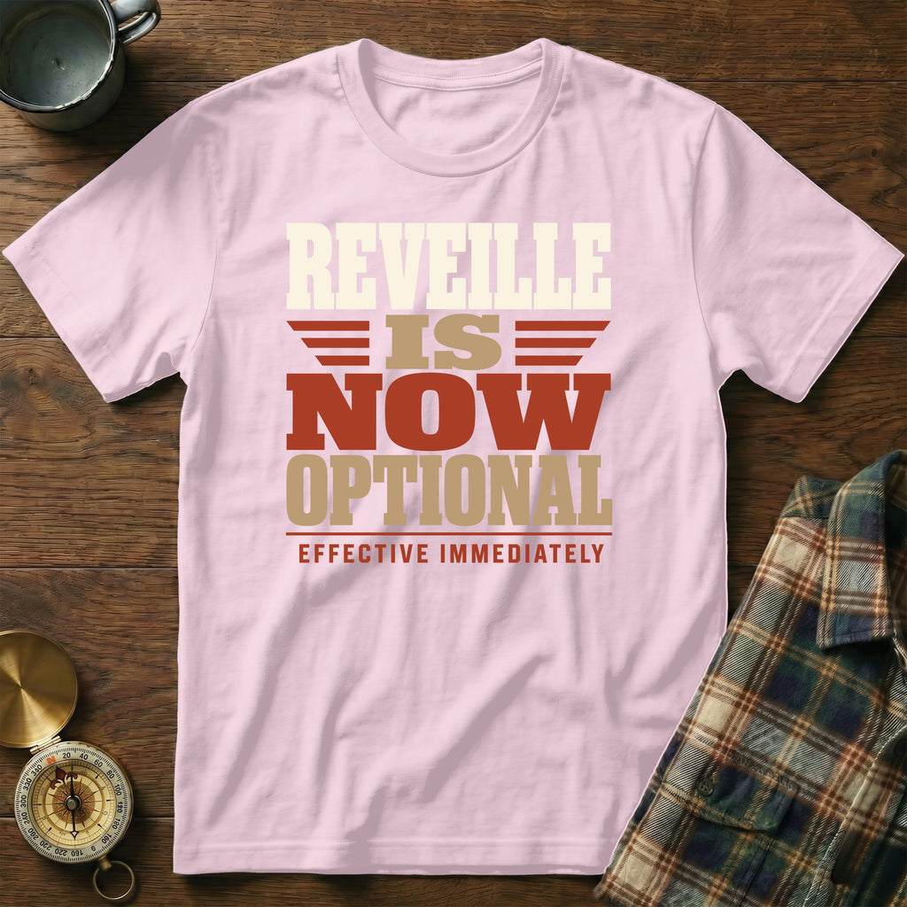 A light pink t-shirt printed with the text "REVELLE IS NOW OPTIONAL EFFECTIVE IMMEDIATELY" lies on a wooden surface beside a mug, a compass, and a plaid shirt.