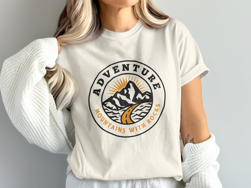 Cream t-shirt with circular design of mountains, road, and sun, text reads "Adventure Mountains With Rocks".