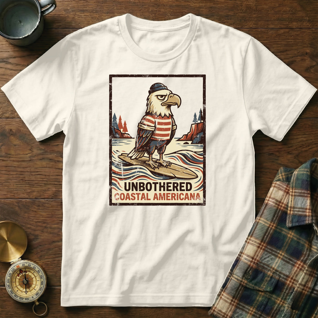 White t-shirt with distressed graphic of a bald eagle in a sailor hat and striped shirt on a surfboard, text "Unbothered Coastal Americana."
