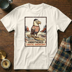 White t-shirt with distressed graphic of a bald eagle in a sailor hat and striped shirt on a surfboard, text "Unbothered Coastal Americana."