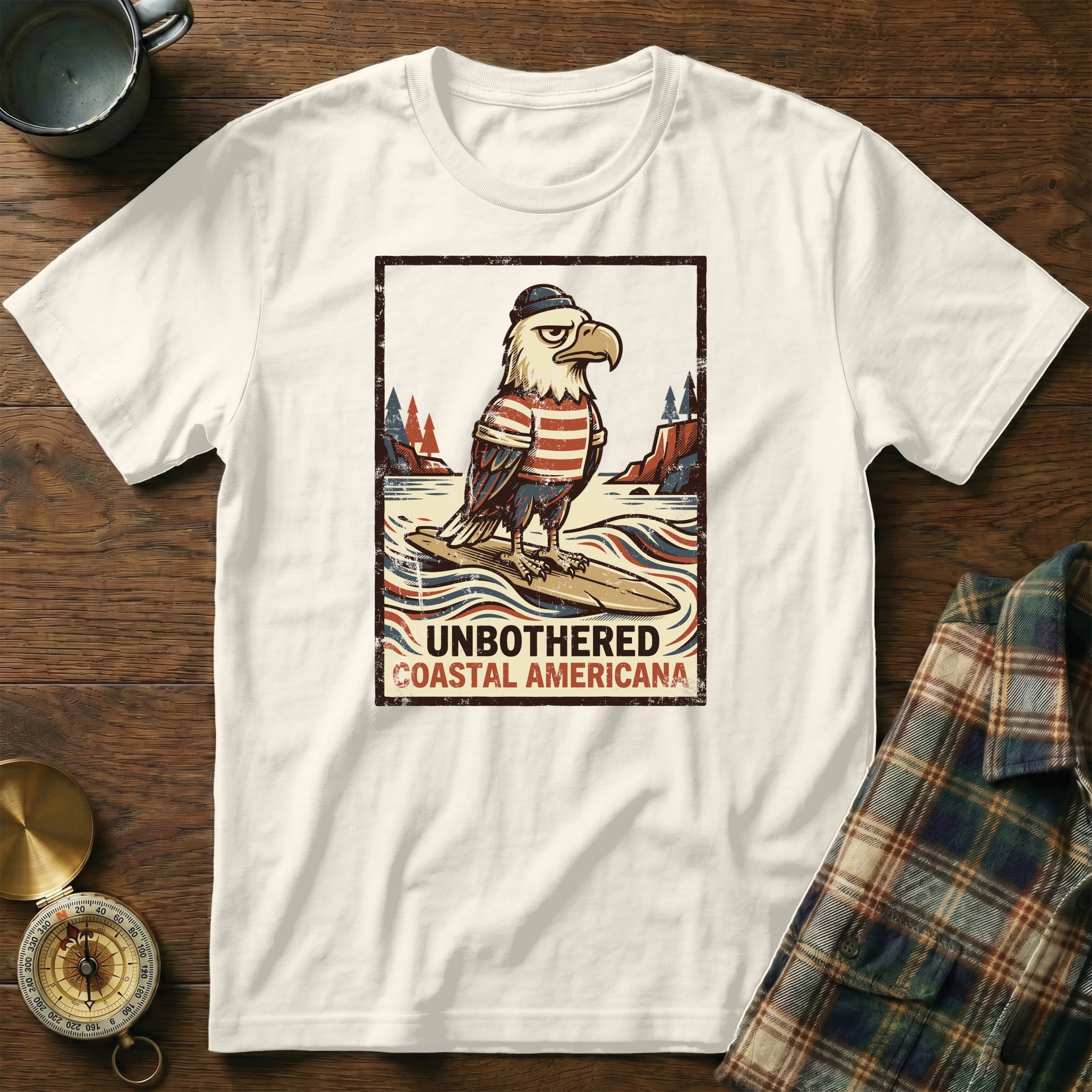 White t-shirt with distressed graphic of a bald eagle in a sailor hat and striped shirt on a surfboard, text "Unbothered Coastal Americana."