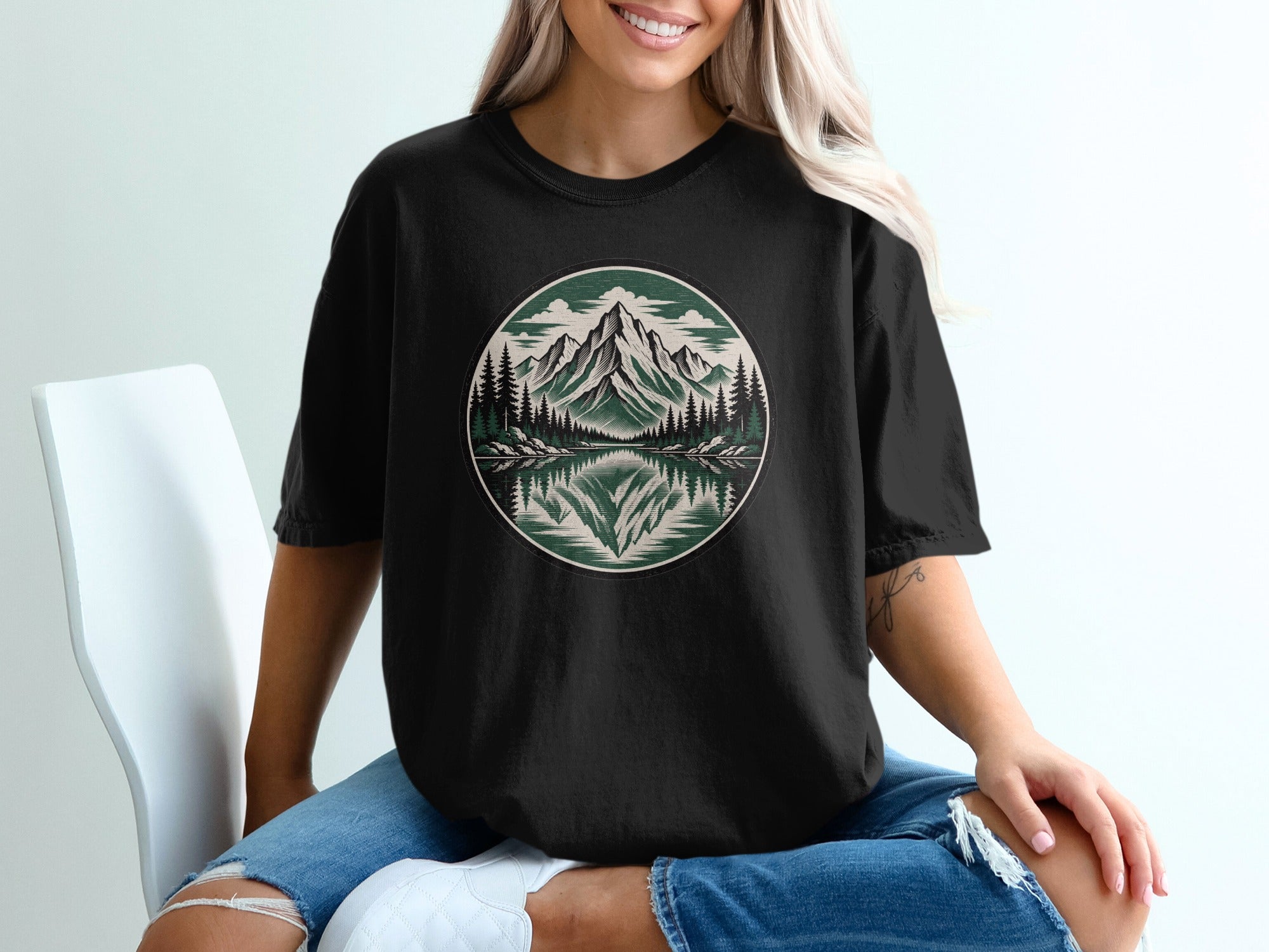 Black t-shirt with a circular mountain and forest scene reflected in water.