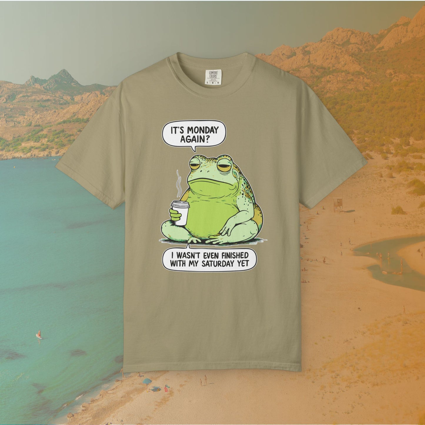 Khaki Tshirt Funny T-shirt graphic featuring a large, grumpy toad holding a to-go coffee cup. The cartoon is captioned with the quotes: "IT'S MONDAY AGAIN? I WASN'T EVEN FINISHED WITH MY SATURDAY YET.