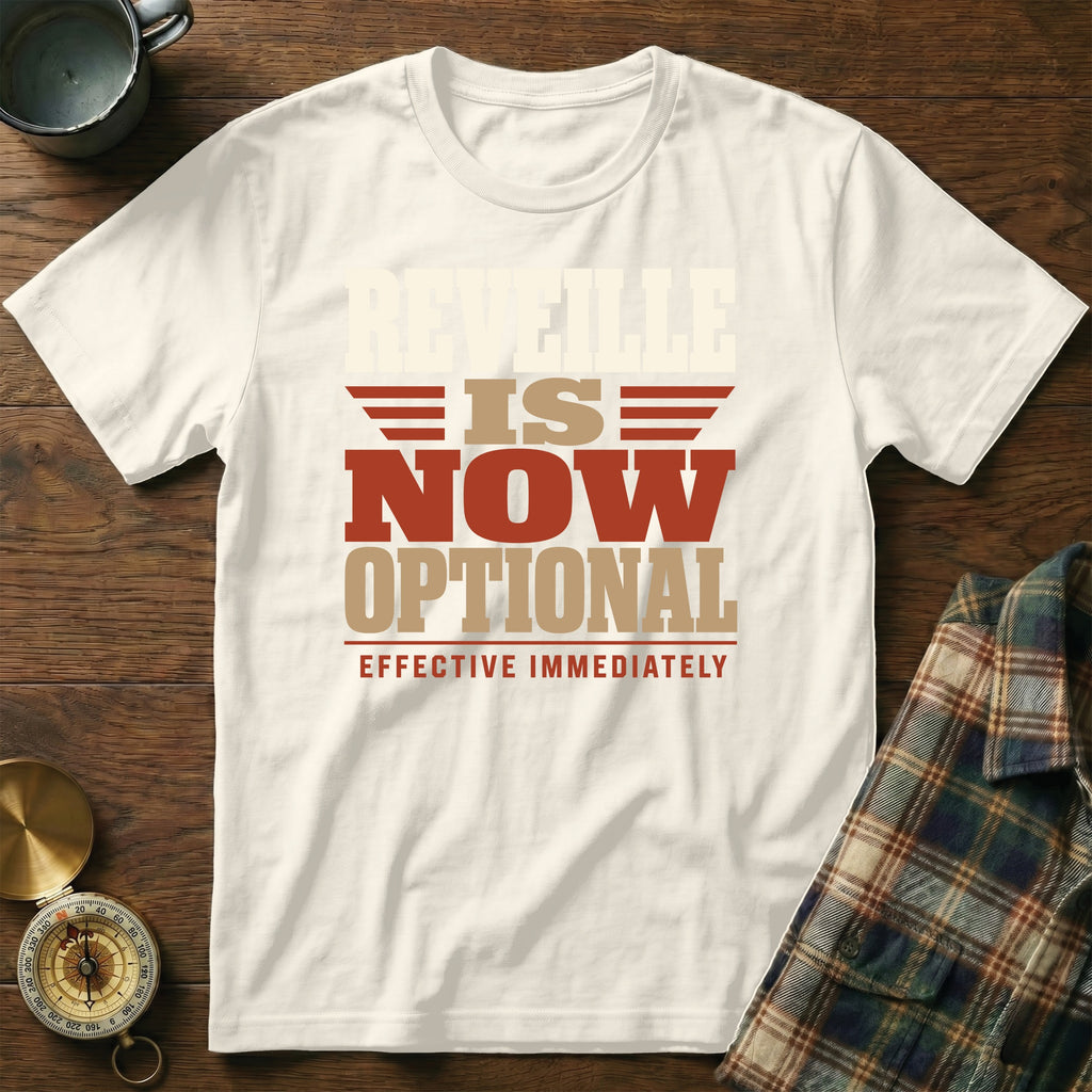 A cream-colored t-shirt with red and tan text reading "Reveille is now optional, effective immediately" on a wooden background.