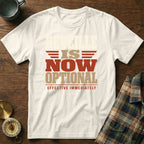 A cream-colored t-shirt with red and tan text reading "Reveille is now optional, effective immediately" on a wooden background.