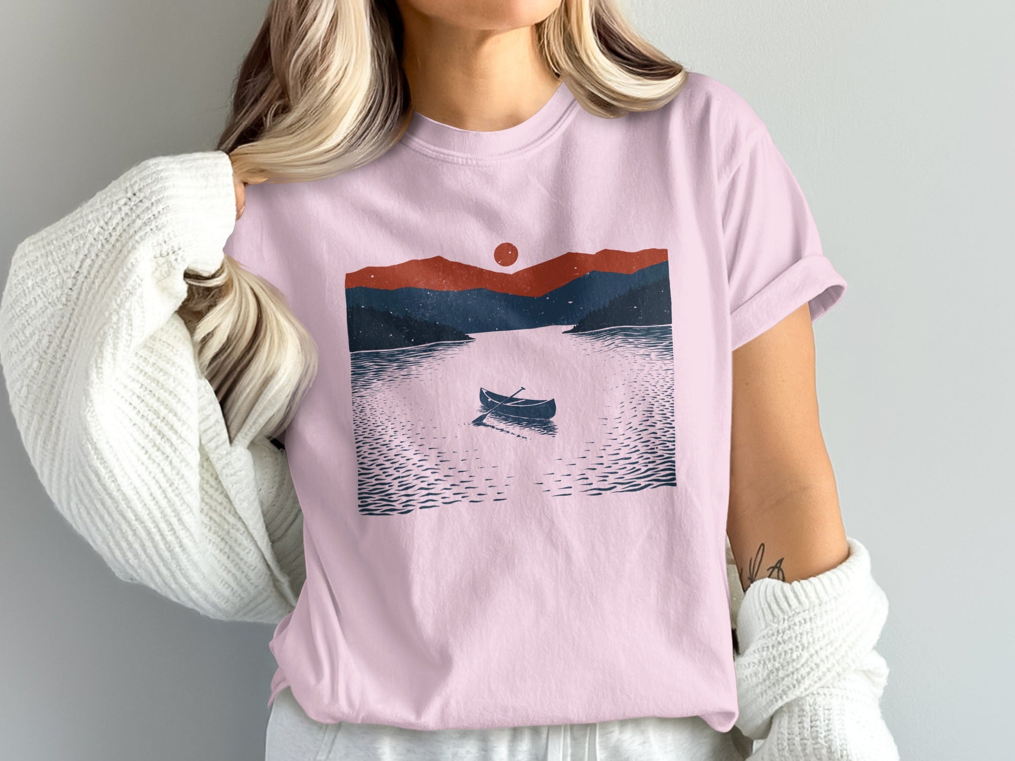 Pink t-shirt with a graphic of a rowboat on a lake at sunset with mountains.