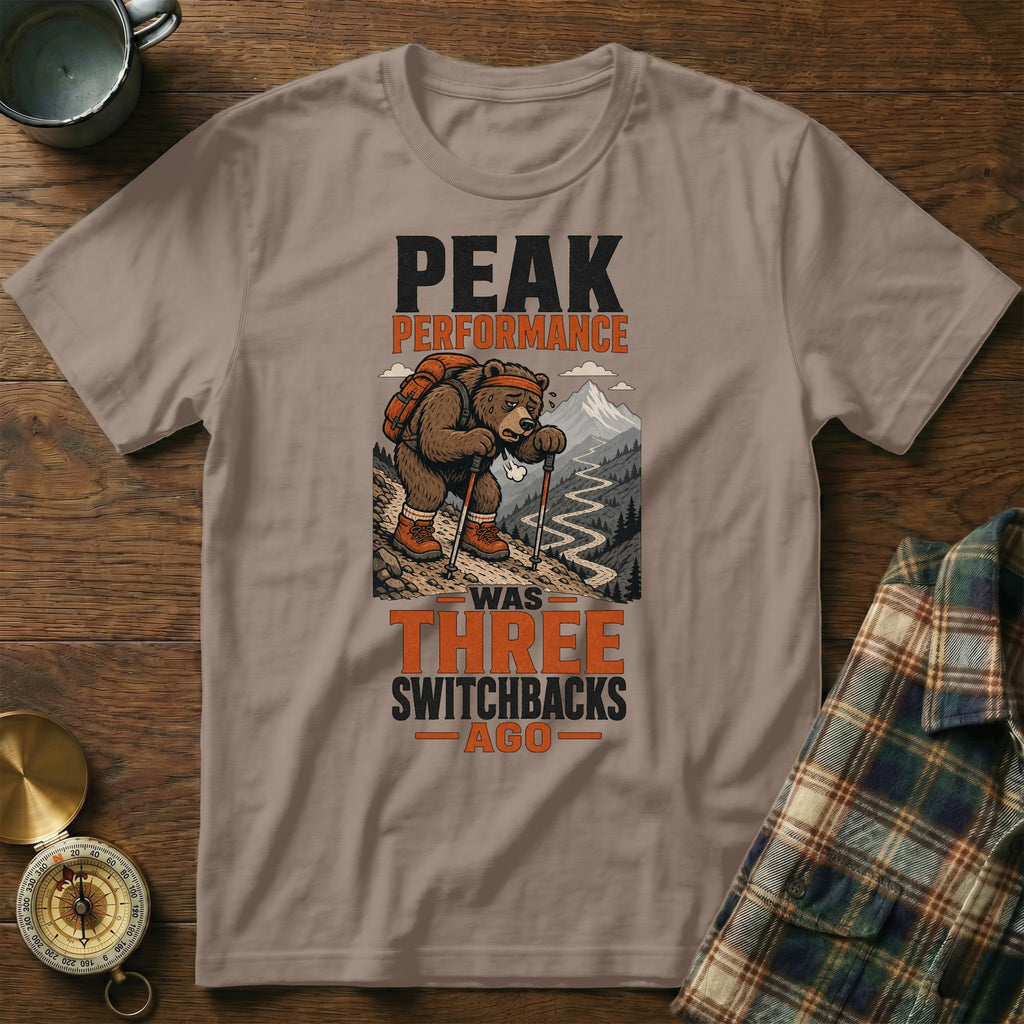 T-shirt graphic with bear hiking uphill, reads "Peak Performance Was Three Switchbacks Ago".