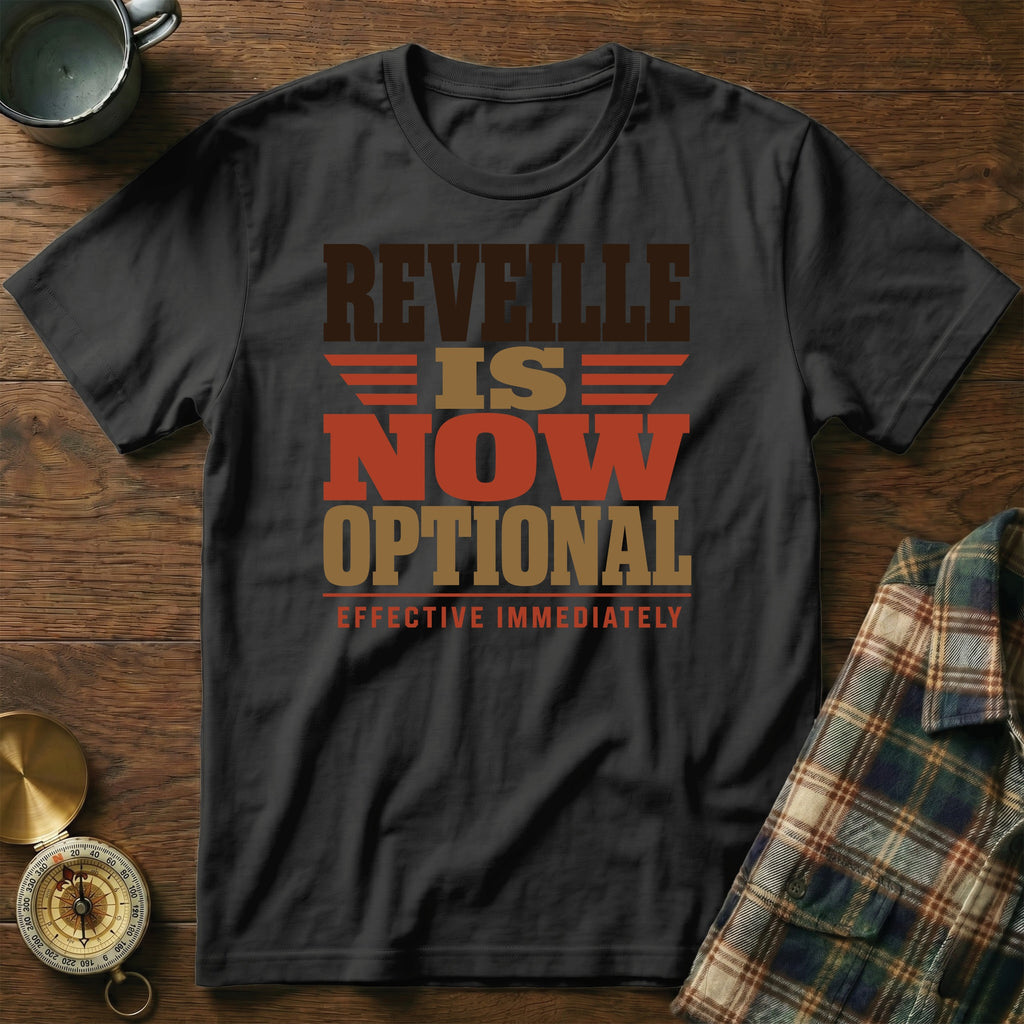 A black t-shirt printed with the text "REVEILLE IS NOW OPTIONAL EFFECTIVE IMMEDIATELY" lies flat on a wooden surface beside a mug, a compass, and a plaid shirt.
