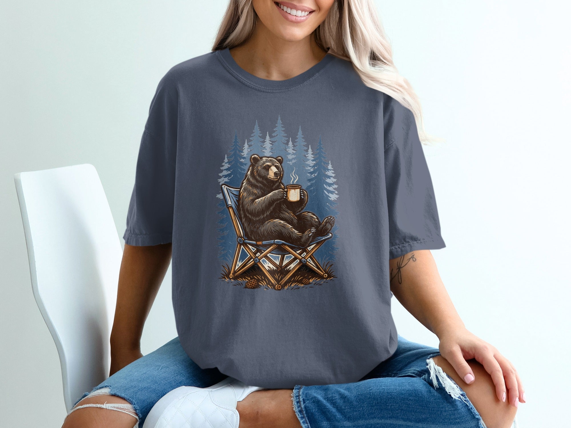 Bear sitting in a camping chair holding a cup by a forest of blue pine trees.