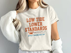 Cream t-shirt with "Low Tide, Lower Standards. Still Showing Up." text and crab graphic.
