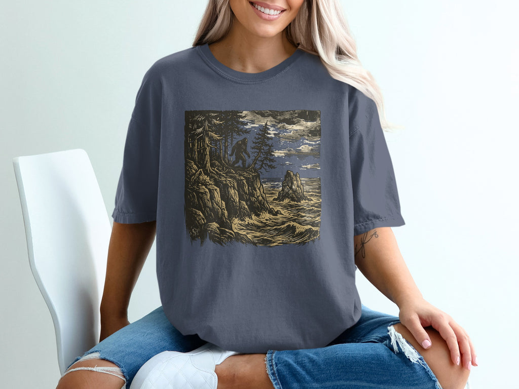 Graphic t-shirt with Bigfoot silhouette on rocky coast overlooking ocean at night.