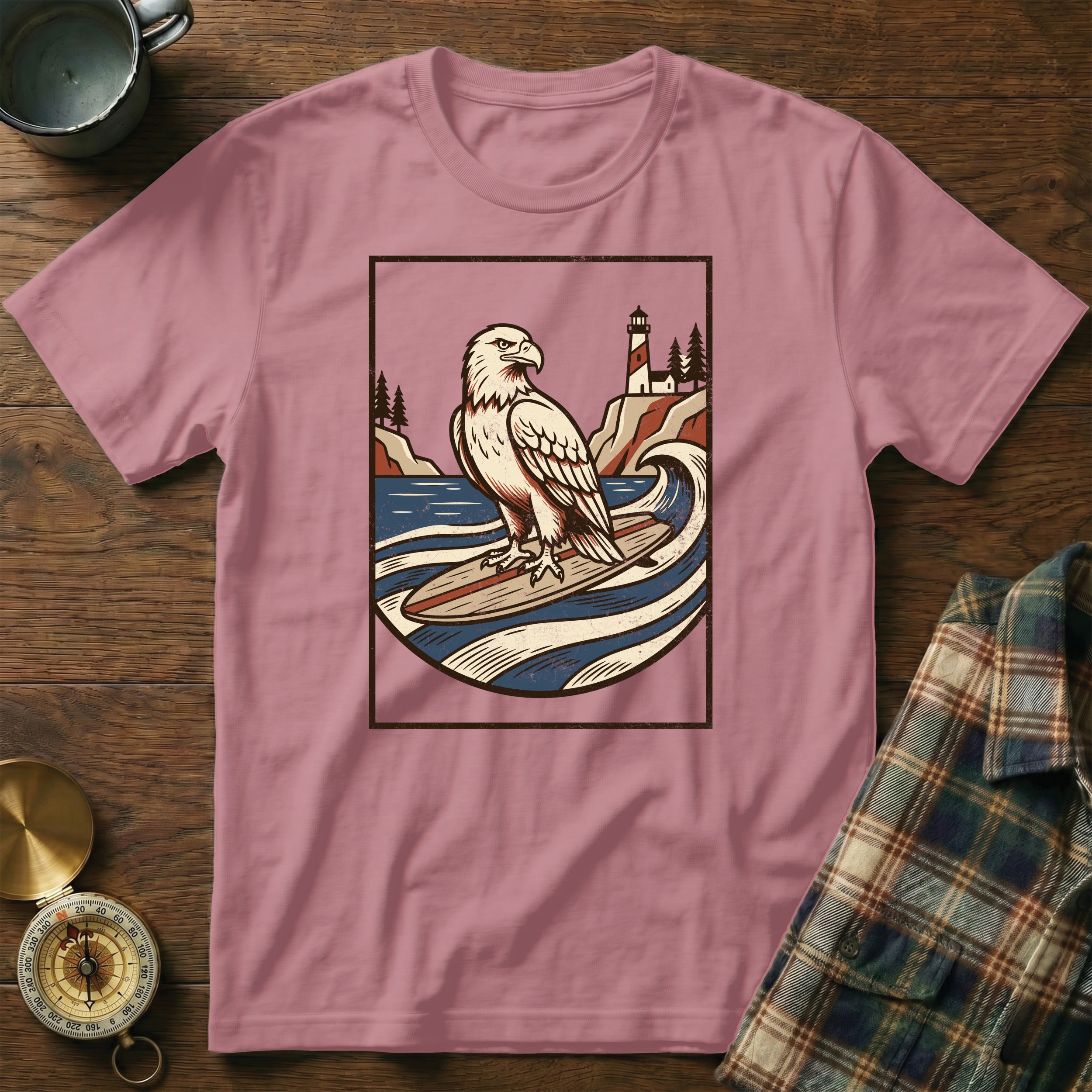 Pink t-shirt with an eagle surfing towards a lighthouse on a rocky shore.