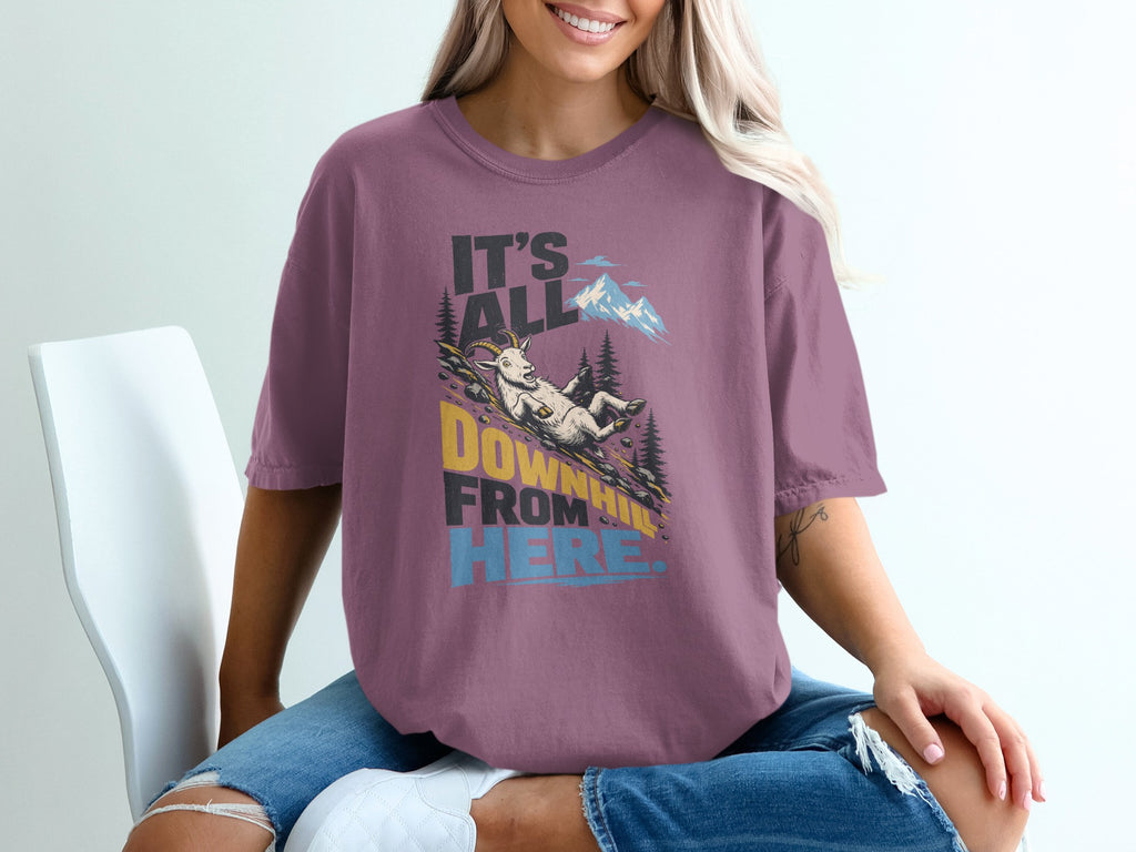 A woman wearing a mauve t-shirt with a graphic of a goat sliding down a mountain and the text it's all downhill from here.