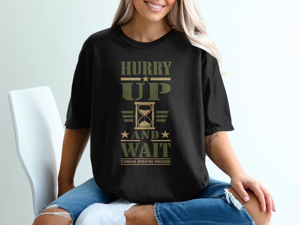 A woman wears a black t-shirt with the green military-style text Hurry Up and Wait.