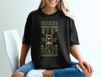 A woman wears a black t-shirt with the green military-style text Hurry Up and Wait.