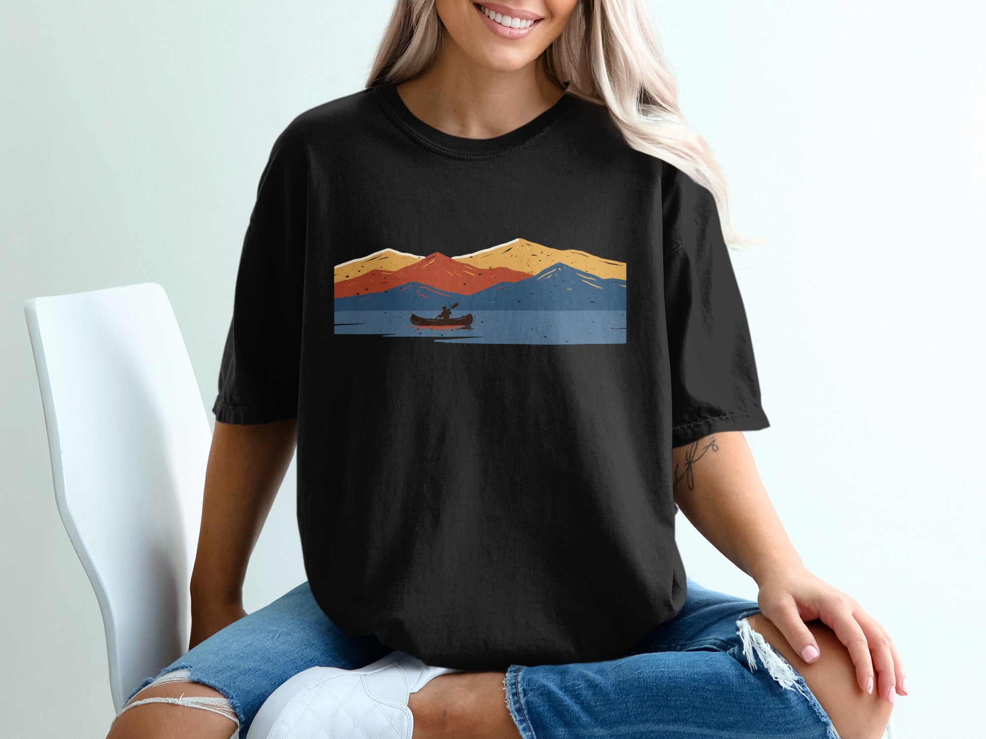 Black t-shirt with a retro graphic of a canoeist on a lake with colorful mountains.