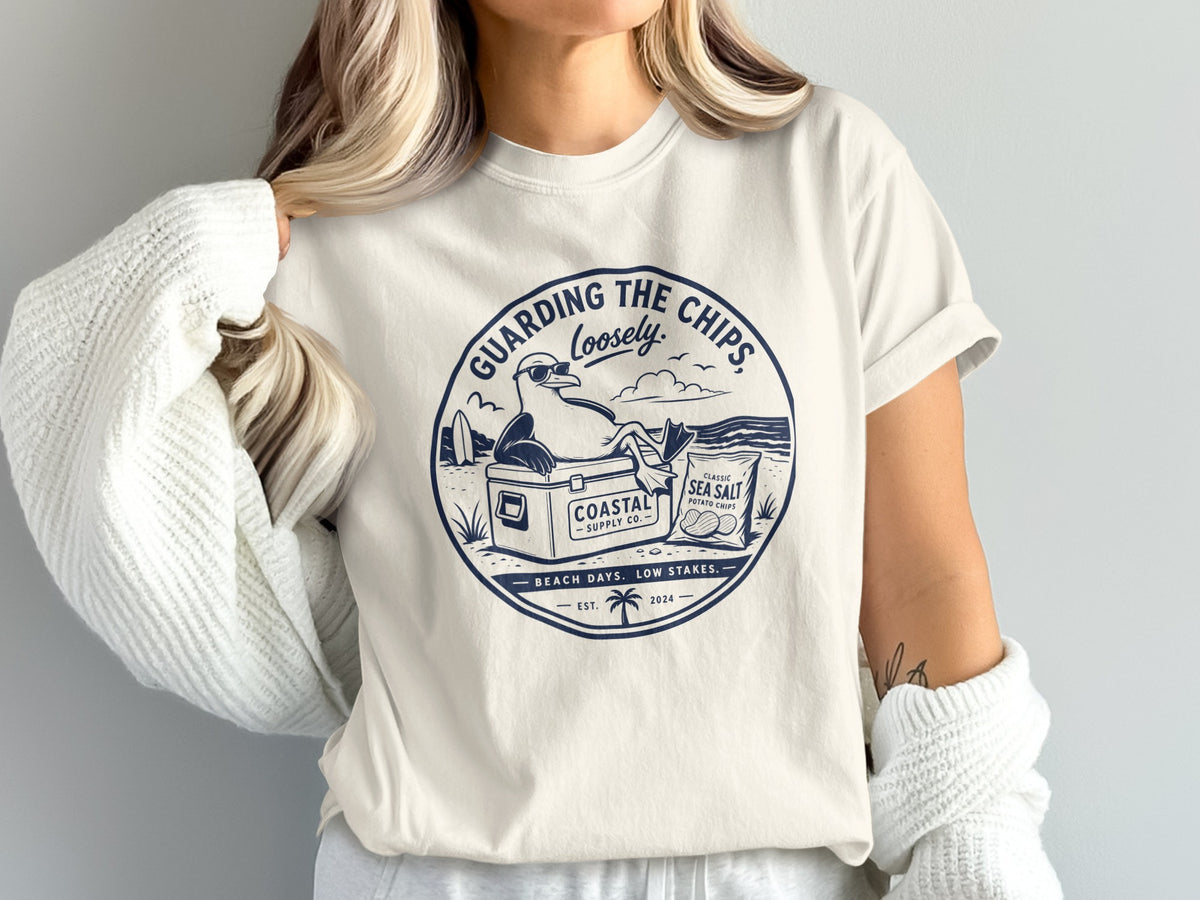 T-shirt graphic with a seagull guarding chips, beach scene, "Guarding the Chips Loosely" text.