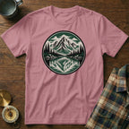 Pink t-shirt with circular mountain and lake graphic, near compass and flannel shirt.