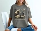 Grey t-shirt with a Japanese-style wave and UFO graphic, text "The Unexplained Tide".