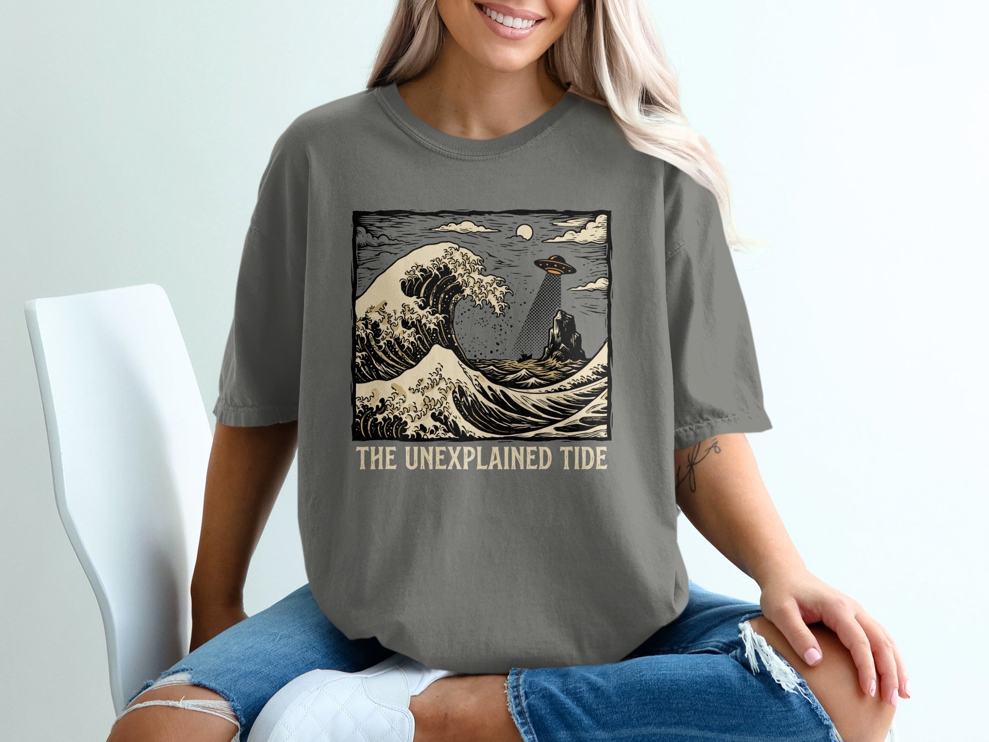 Grey t-shirt with a Japanese-style wave and UFO graphic, text "The Unexplained Tide".