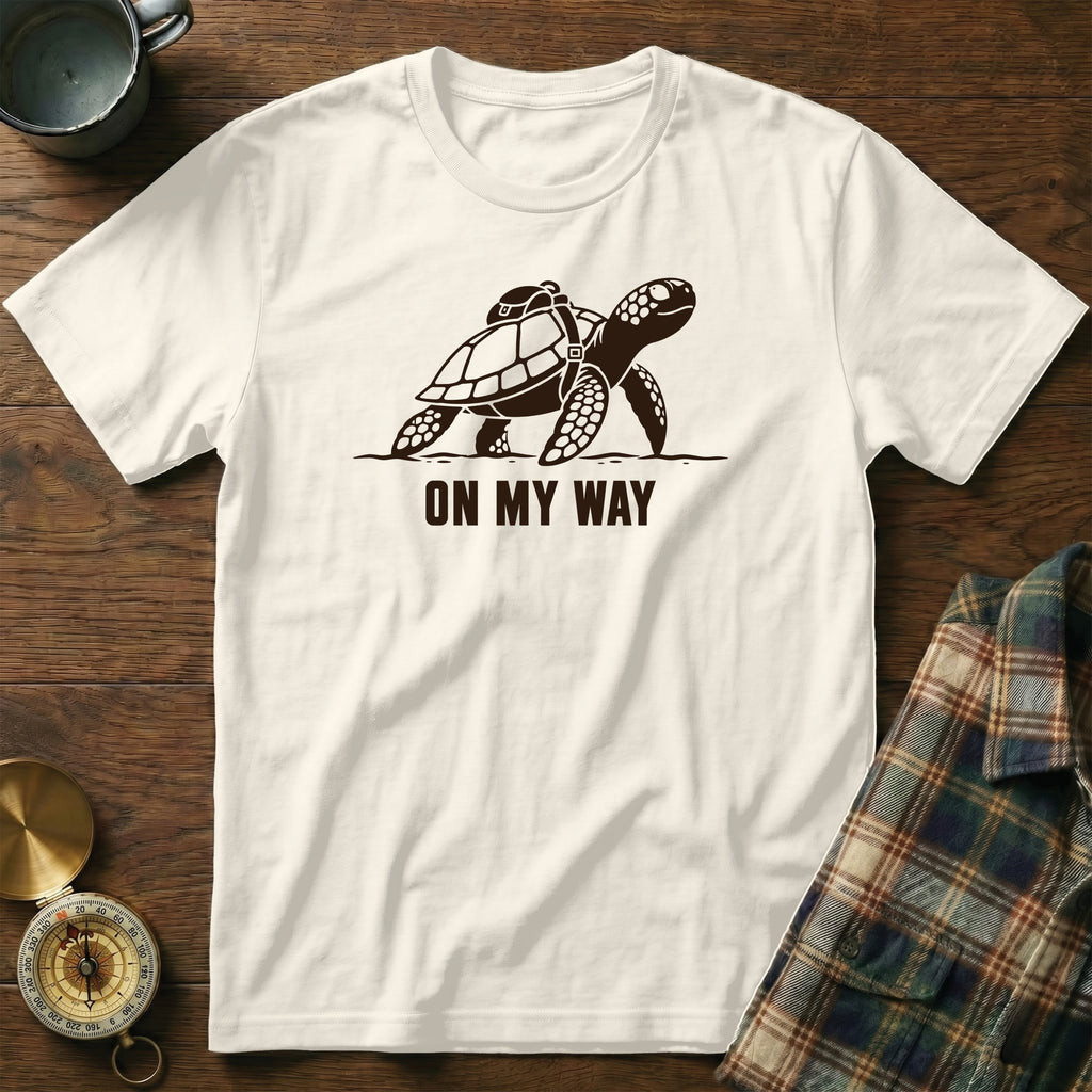 White t-shirt with a dark brown graphic of a turtle wearing a backpack and the text "ON MY WAY".