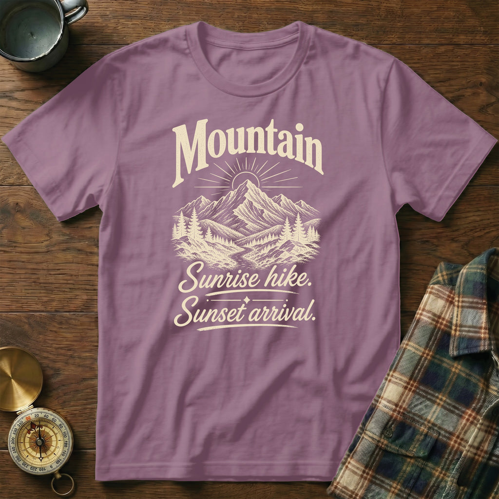 Dusty purple t-shirt featuring a mountain graphic and the text Mountain Sunrise hike. Sunset arrival. on a wooden surface.