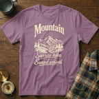 Dusty purple t-shirt featuring a mountain graphic and the text Mountain Sunrise hike. Sunset arrival. on a wooden surface.