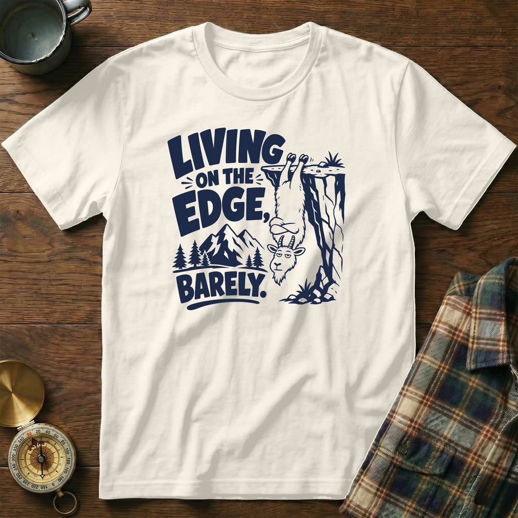 White t-shirt with navy graphic of goat hanging from cliff, text "LIVING ON THE EDGE, BARELY."
