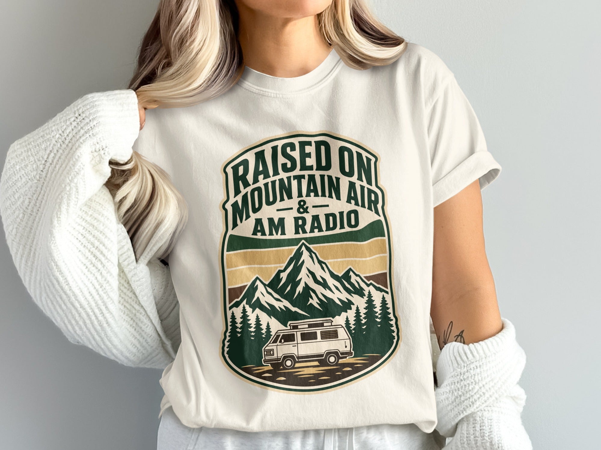White t-shirt with a vintage crest graphic of mountains, trees, and a van. Text reads "Raised on Mountain Air & AM Radio".