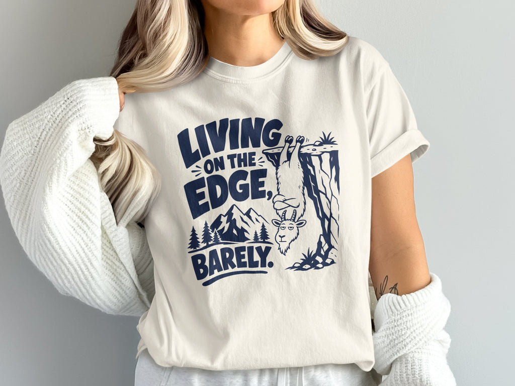 T-shirt with navy graphic: goat hanging from cliff with text "LIVING ON THE EDGE, BARELY."