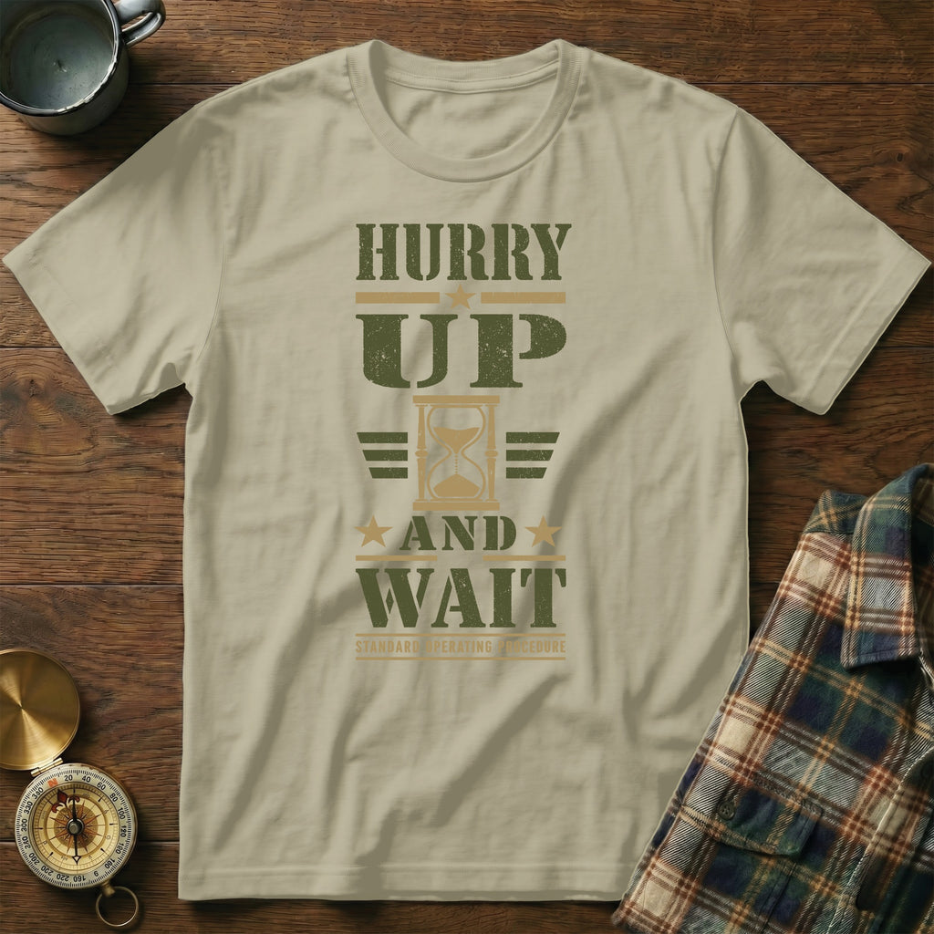 A light green t-shirt with green distressed text reading HURRY UP AND WAIT centered on a wooden surface.