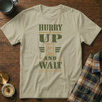 A light green t-shirt with green distressed text reading HURRY UP AND WAIT centered on a wooden surface.