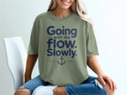 A blonde woman wearing an olive green t-shirt with navy blue text that reads Going with the flow. Slowly. and an anchor graphic.