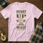 A light pink t-shirt with olive green text reading "Hurry Up and Wait" and an hourglass graphic.