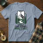 Light blue t-shirt with a screen print of a mountain landscape within a phone outline and text "LESS SCROLL. MORE STROLL."