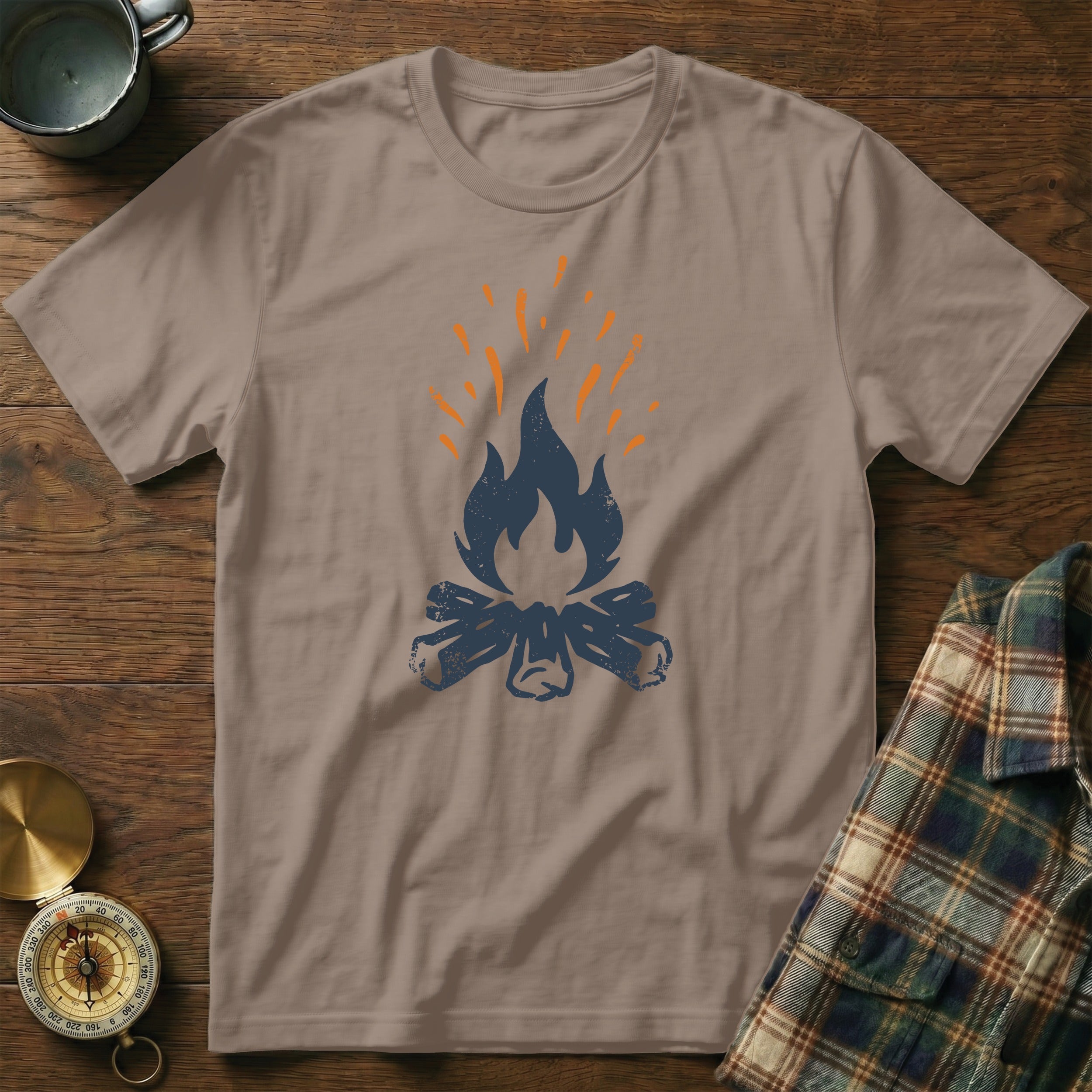 Campfire graphic on a taupe t-shirt, laid on a wooden surface with a compass and plaid shirt.