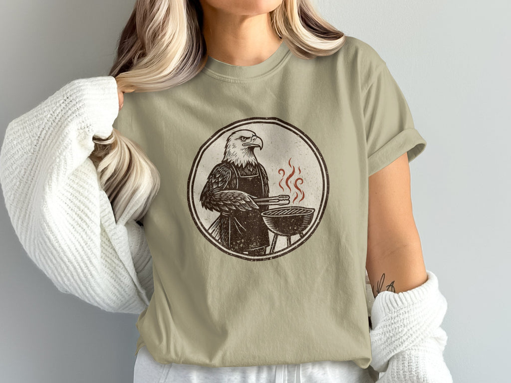 Eagle in apron grilling on a barbecue, brown graphic on a tan t-shirt.