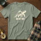 Olive green t-shirt with a graphic of a sea turtle wearing a backpack and text "ON MY WAY".