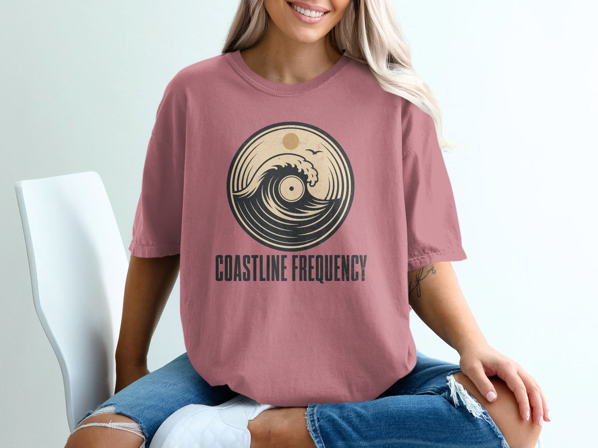Dusty rose t-shirt with circular wave and sun graphic and "Coastline Frequency" text.