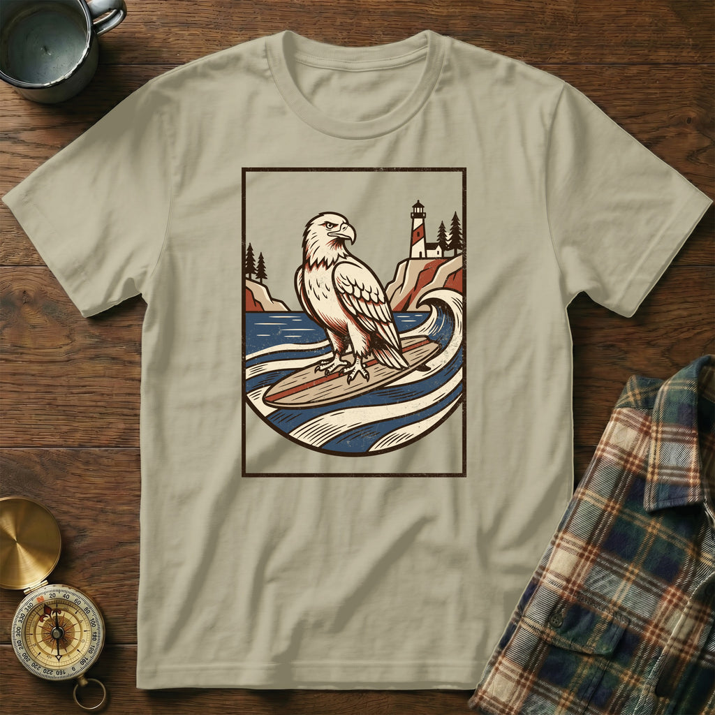 Eagle surfing on a surfboard with lighthouse and ocean graphic on t-shirt.