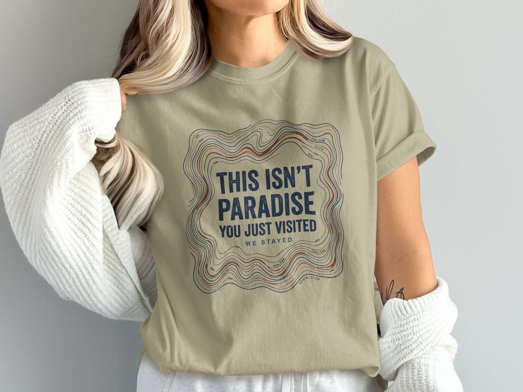 Tan t-shirt with topographic lines and text "THIS ISN'T PARADISE YOU JUST VISITED WE STAYED".