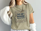 Tan t-shirt with topographic lines and text "THIS ISN'T PARADISE YOU JUST VISITED WE STAYED".