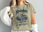 A person wears a beige t-shirt featuring a blue mountain graphic and the text Mountain Sunrise hike Sunset arrival.