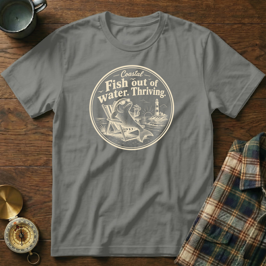 A grey crew-neck t-shirt featuring a circular graphic of a fish in a beach chair holding a drink with the text Coastal Fish out of water. Thriving.