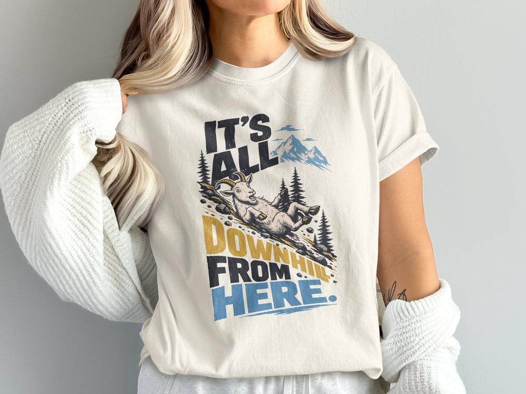 A cream-colored t-shirt with a graphic of a fainting goat sliding down a hill, accompanied by the text It's all downhill from here.